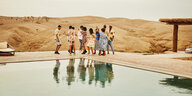 Menschen in Shorts und Kleidern tanzen an einem Pool vor einer Wüstenlandschaft