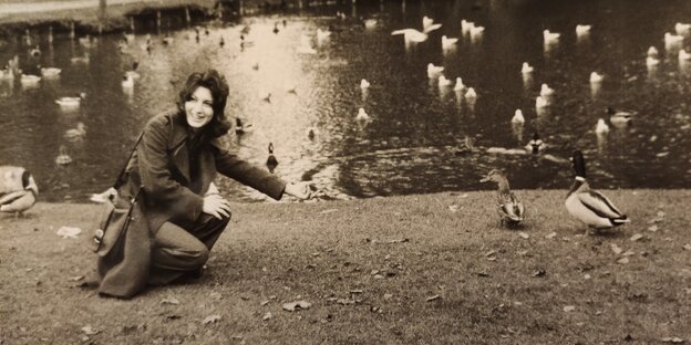 Schwarz-weiß-Foto: eine lächelnde dunkelhaarige junge Frau hockt inmitten von Enten an einem Teich