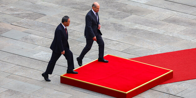 Der chinesische Premierminister und Olaf Scholz gehen auf ein mit rotem Teppich überzogenes Podest