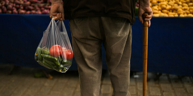 Die Beine eines Mannes, der einen Gehstock in der einen und eine Plastiktüte mit Gemüse in der anderen hält.