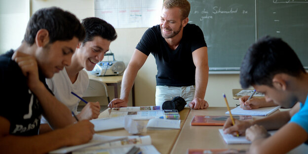 Drei Schüler sitzen an einem Tisch, ein Lehrer steht vor dem Tisch