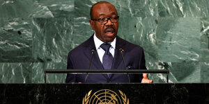 Ali Gabon steht an einem Rednerpult, er trägt eine schwarze Brille und einen Anzug