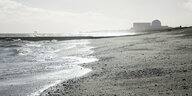 Wellen ziehen Schaumkronen über einen Sandstrand, im Hintergrund ist einr Atomkraftanlage zu erkennen