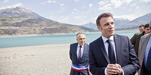 Präsident Macron m Anzug am Strand. Andere Männer im Anzug stehen um ihn.