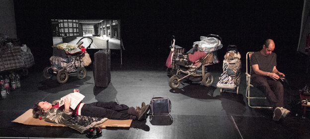 Eine Schauspielerin liegt umgeben von Kinderwagen und Koffern auf der Bühne. Ein Mann sitzt daneben auf einem Stuhl Eine Schauspielerin liegt umgeben von Kinderwagen und Koffern auf der Bühne. Ein Mann sitzt daneben auf einem Stuhl