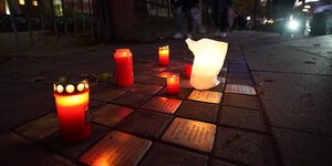 Kerzen stehen zwischen den goldenen Stolpersteinen, die in der Straße eingelassen sind. Menschen gehen im Hintergrund die Straße entlang.