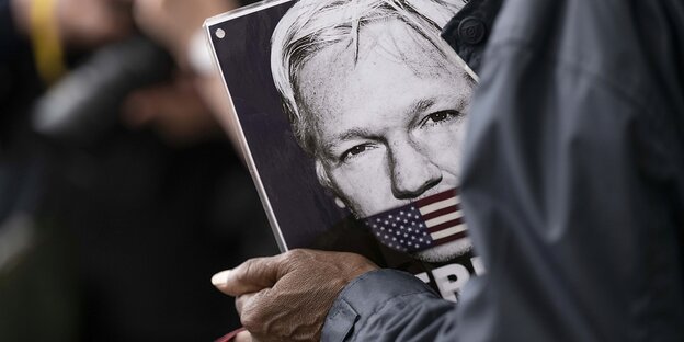 Jamns hält ein Foto von Julan Assange in den Händen. SEin Mund ist mit einer US-Fahne zugebunden