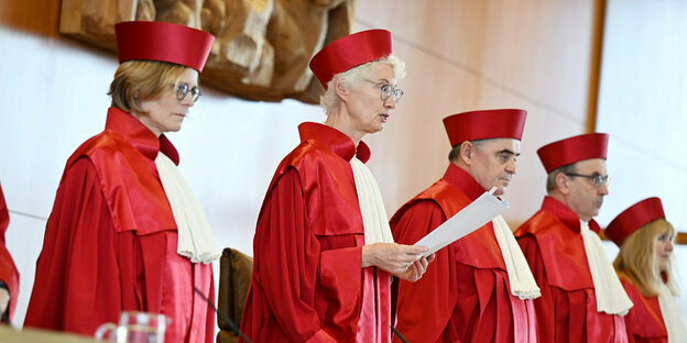 Fünf Richter*innen in roten Roben und rotem Hut stehen bei der Urteilsverkündung. Astrid Wallrabenstein liest das Urteil vor