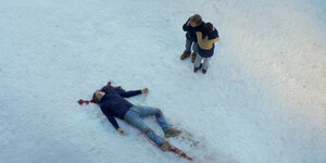 Blick von oben auf einen Mann, der im Schnee liegt, mit blutigen Spuren. Zwei Personen stehen daneben