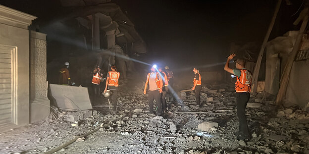 Männer in orangenen Westen und mit Lampen vor zerstörten Gebäuden bei Nacht