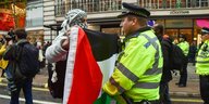 ein mit einem Palästinensertuch vermummter Mann hält einem Polizisten, eine Palästinaflagge entgegen