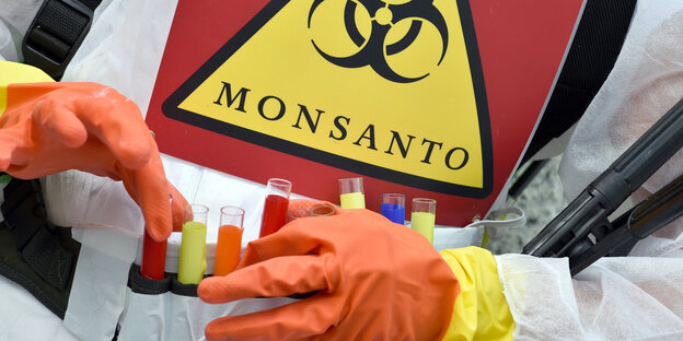 Handschuhe fassen Röhrchen mit buntem Pulver an, Schild mit der Aufschrift Monsanto Handschuhe fassen Röhrchen mit buntem Pulver an, Schild mit der Aufschrift Monsanto