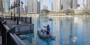Ein Mann fischt mit einem kleinen Fischernetzt etwas aus einem künstlichen See, im Hintergrund sind sehr hohe, moderne Hochhäuser