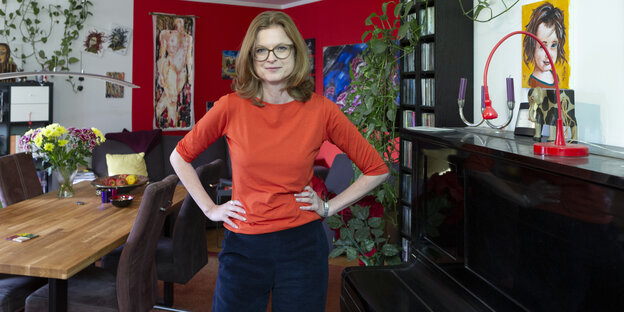 Eine Frau mittleren Alters mit Brille und orangenem Oberteil steht in einer Wohnung mit bunten Wänden und vielen Bildern an den Wänden
