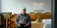 Wolfgang Seibert sitzt auf einer Bank in einem Gebetsraum, vor ihm ein Kerzenständer mit sieben angezündeten Kerzen