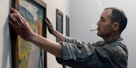 Ein Mann im Kittel und mit Zigarette im Mund rückt ein Gemälde auf einer Wand zurecht