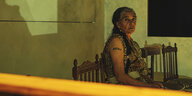 Eine alte Frau mit Tätowierung am Oberarm sitzt auf einem Stuhl und schaut von der Seite aus in die Kamera