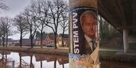 An einer Brückensäule klebt ein beschädigtes Wahlplakat von Geert Wilders, im Hintergrund sind ein gut gefüllter Kanal, Bäume und rote Klinkerhäuser zu sehen.