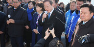 Ein Mann mit blauer Krawatte ist von einer Menschenmenge umrignt und spricht in ein Mikrofon