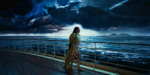 Eine Frau mit langen schwarzen Haaren an der Reling eines Schiffs. Im Hintergrund eine Insel am Horizont schwarze, fantastisch übertrieben ausehende Wolken