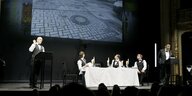 Theaterbühne mit einer Leinwand, die eine Straße zeigt, Darsteller im Kellnerdress sitzen und stehen an einem weiß eigedeckten Tisch