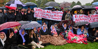 JesidInnen sitzen und stehen mit Regenschirmen und Transparenten auf dem Rasen und fordern einen Stopp der Abschiebungen in den Irak