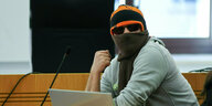 Einer der Angeklagten sitzt vermummt mit Mütze und dunkler Sonnenbrille im Gerichtssaal