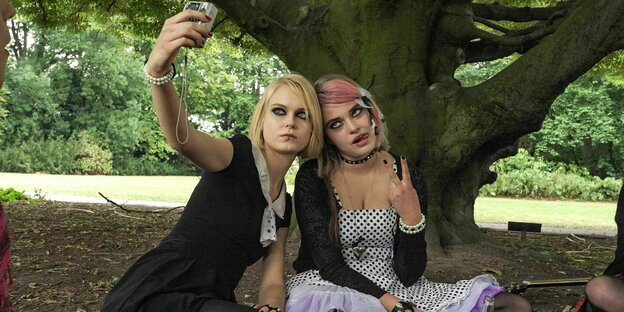 Zwei gestylte Mädchen sitzen unter einem Baum im Park und machen ein Selfie