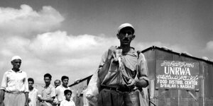 Ein Schwarz-Weiß-Foto zeigt Menschen vor einem Holzschuppen mit der Aufschrift "UNRWA"