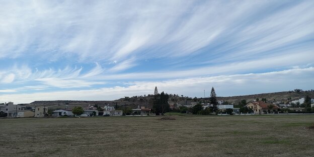 Panoramaansicht eines kleinen Ortes, im Vordergrund Felder, darüber wolkig-blauer Himmel