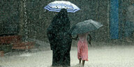 Eine Frau und ein Kind laufen mit Regenschirmen durch Starkregen über eine überflutete Straße. Die frau trägt einen Tschador