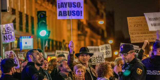 Demonstrierende Menge, darüber auf einem Schild wird der Rücktritt von Ayuso gefordert