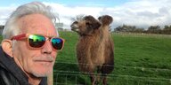 Johnny Dowd steht neben einem Kamel, der Himmel ist bewölkt ,er trägt einer verspiegelte Sonnenbrille