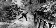 Soldaten rennen sich duckend über eine unbefestigte Straße, vorne steht ein Panzer, hinteer ihnen liegt ein Lastwagen im Graben