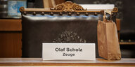 Auf einem Schild auf einem Tisch steht: Olaf Scholz, Zeuge