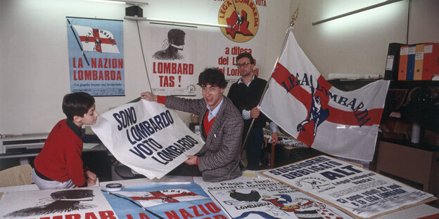 De jonge Umberto Bossi zit over Plakaten der Lega Nord an einem Tisch