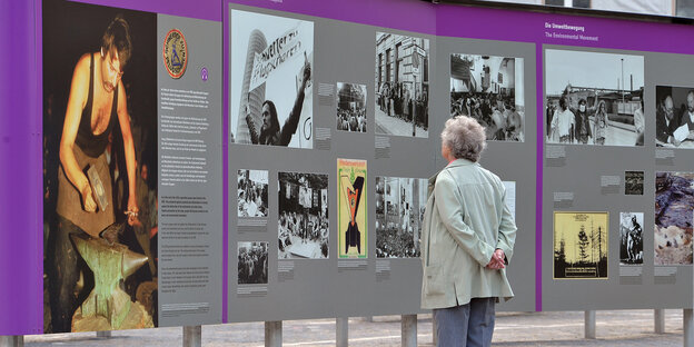 Eine ältere Frau liest sich die Texte einer Tafel der Ausstellung "Revolution und Mauerfall" durch. Sie steht mit dem Rücken zur Kamera. Die Tafel ist lilafarben unterlegt, sie zeigt das Foto eines Schmieds und Informationstexte.