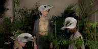 Drei Menschen mit Vogelmasken stehen zwischen Grünpflanzen