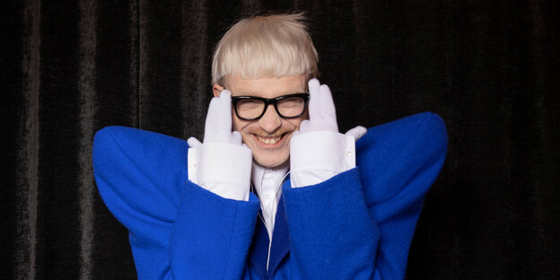Joost Klein mit überschnittenen hochgestelltem grell blauem Schulterpolster hebt die Finger zu den Augen und trägt weiße Handschuhe
