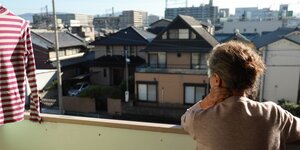 Blick von einem Balkon. Eine ältere Frau steht mit dem Rücken zur Kamera und blick von ihrem Balkon aus auf eine Stadt. Sie hat das Gesicht auf die linnke Hand gestützt. Links im Bild hängt ein rot-weiß gestreiftes Hemd auf einem Bügel zum Trocknen in der Sonne.