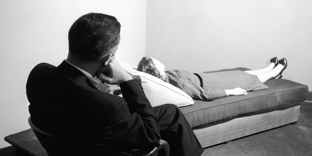 Eine Frau liegt ausgestreckt auf einem Sofa, hinter ihr sitzt ein Mann in einem Sessel