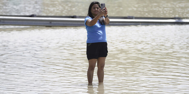 Eine Frau steht auf einer knöcheltief überfluteten Straße und macht ein Selfie