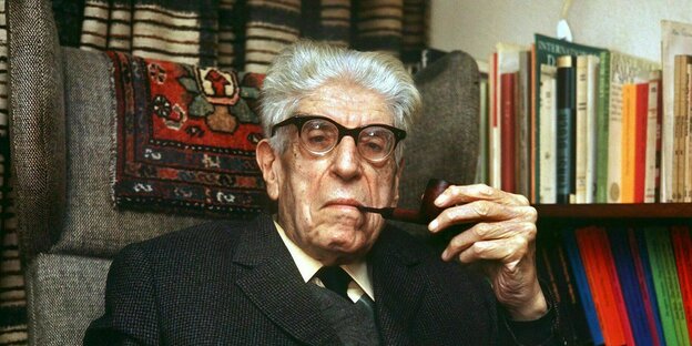 Ernst Bloch, mit grauen Haaren, sitzt in seinem Sessel vor einer Bücherwand und raucht eine Pfeife