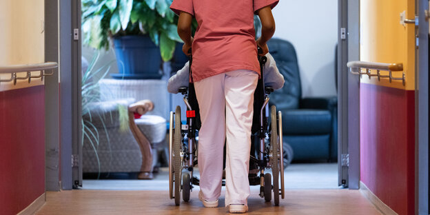 Ein Flur in einem Pflegeheim. Ein*e Pfleger*in ist von hinten zu sehen, wie sie eine*n Bewohner*in in einem Rollstuhl schiebt.