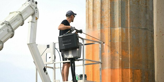 Ein Mann steht auf einer Hebebühne und reinigt mit einem Hochdruckreiniger einen mit oranger Farbe besprühten Pfeiler des Brandenburger Tors