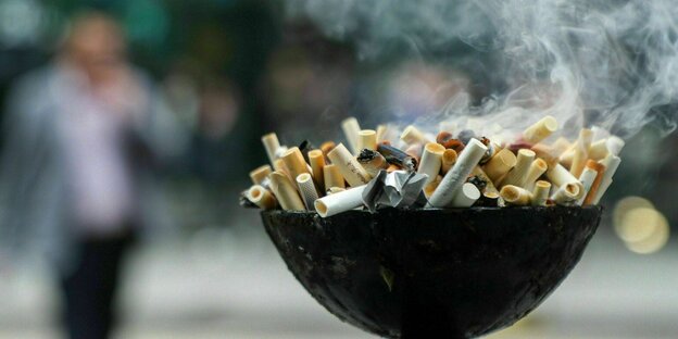 Ein großer, überquellender Aschenbecher mit rauchenden Zigarettenstummeln