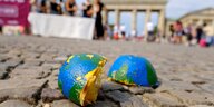 Eine erdenähnlich blau-grün bemalte Orange liegt halbiert und ausgepresst auf dem Boden vor dem Brandenburger Tor in Berlin