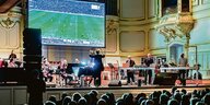 Das Ensemble Resonanz und die Band des Elektronik-Künstlers Matthew Herbert stehen auf der Bühne der Laiszhalle. Im Hintergrund ist ein Bildschirm, auf dem das Fußballspiel übertragen wird.