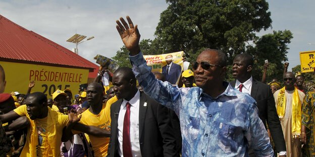 Mann in blauem Hemd mit Sonnenbrille winkt, neben ihm ein Mann in Anzug, dahinter viele in gelben T-Shirts