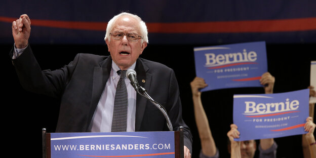 Ein weißhaariger Mann spricht von einem Pult. Im Hintergrund Bernie-Plakate Ein weißhaariger Mann spricht von einem Pult. Im Hintergrund Bernie-Plakate